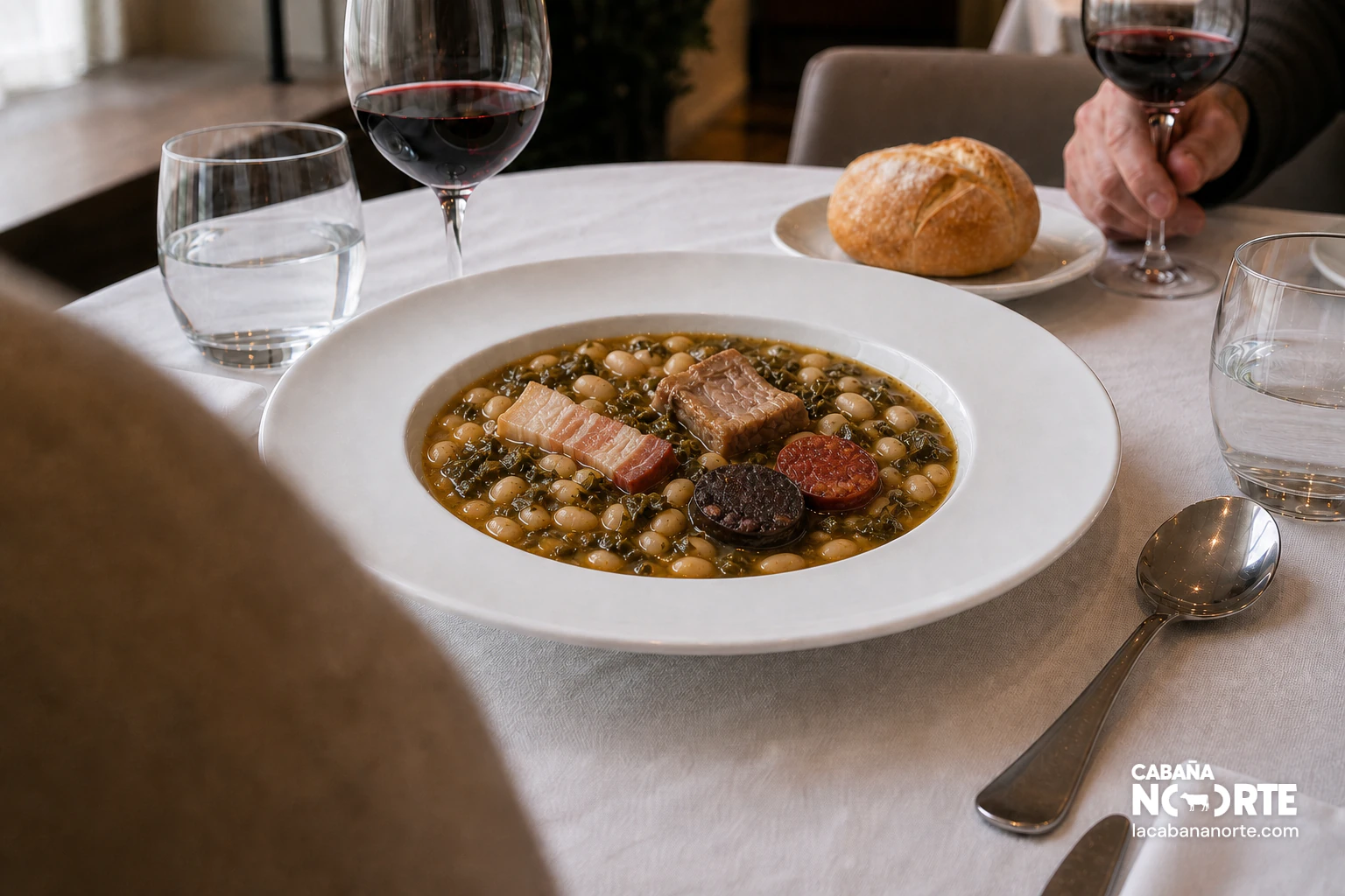 Plato de cocido montañés servido en un restaurante con pan, vino tinto y compango sobre mantel blanco