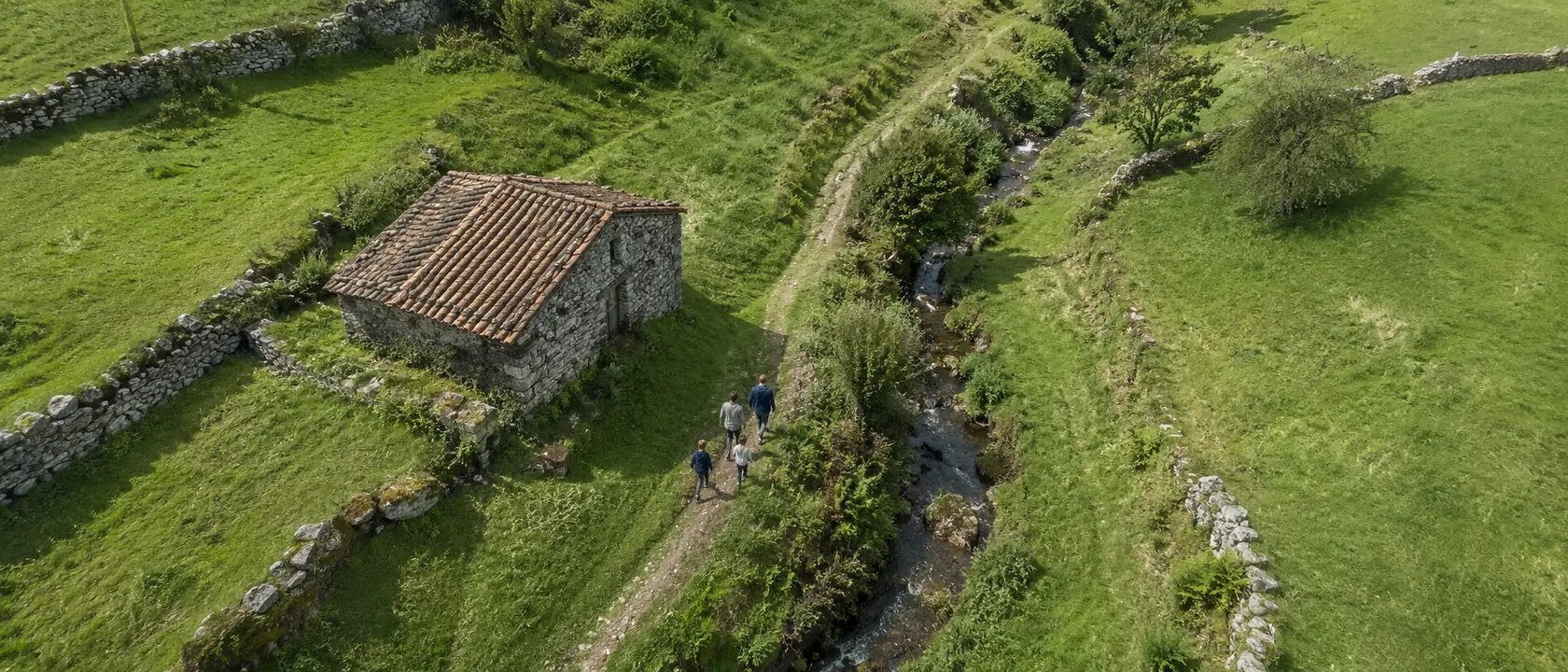 Familia caminando junto a un riachuelo y una cabaña pasiega en los Valles Pasiegos, Cantabria