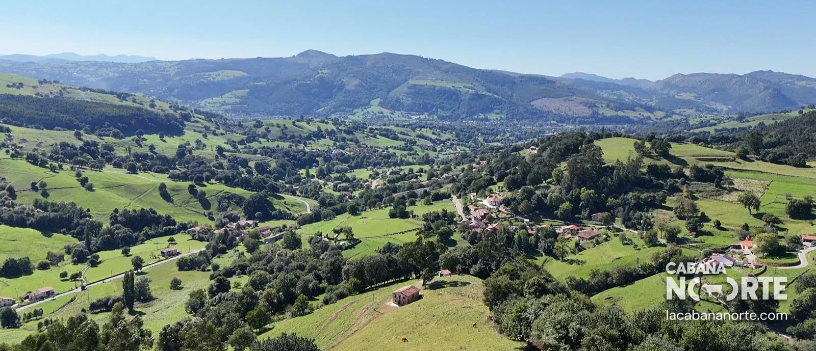 Vista panorámica de un valle verde con prados, casas dispersas y montañas bajo un cielo despejado.